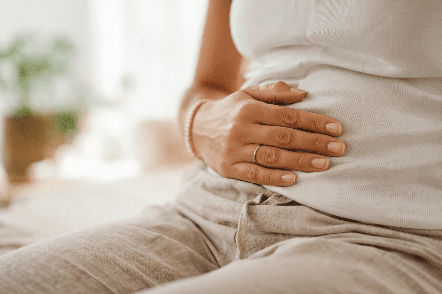 Close-up of a woman’s abdomen with her hand gently resting on her stomach, soft natural lighting, neutral beige clothing, and a blurred white background creating a calm and introspective atmosphere.