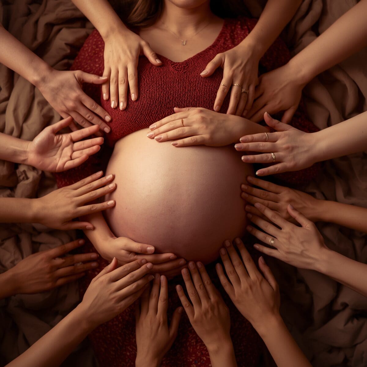 a pregnant woman holding her belly with many supportive hands reaching out toward her. The face isn't visible in any of them, focusing instead on the belly and the circle of caring hands creating a warm, nurturing atmosphere of community support